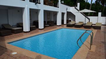 Piscina con sillas de descanso y sombrillas, rodeada por un edificio blanco con varios balcones.
