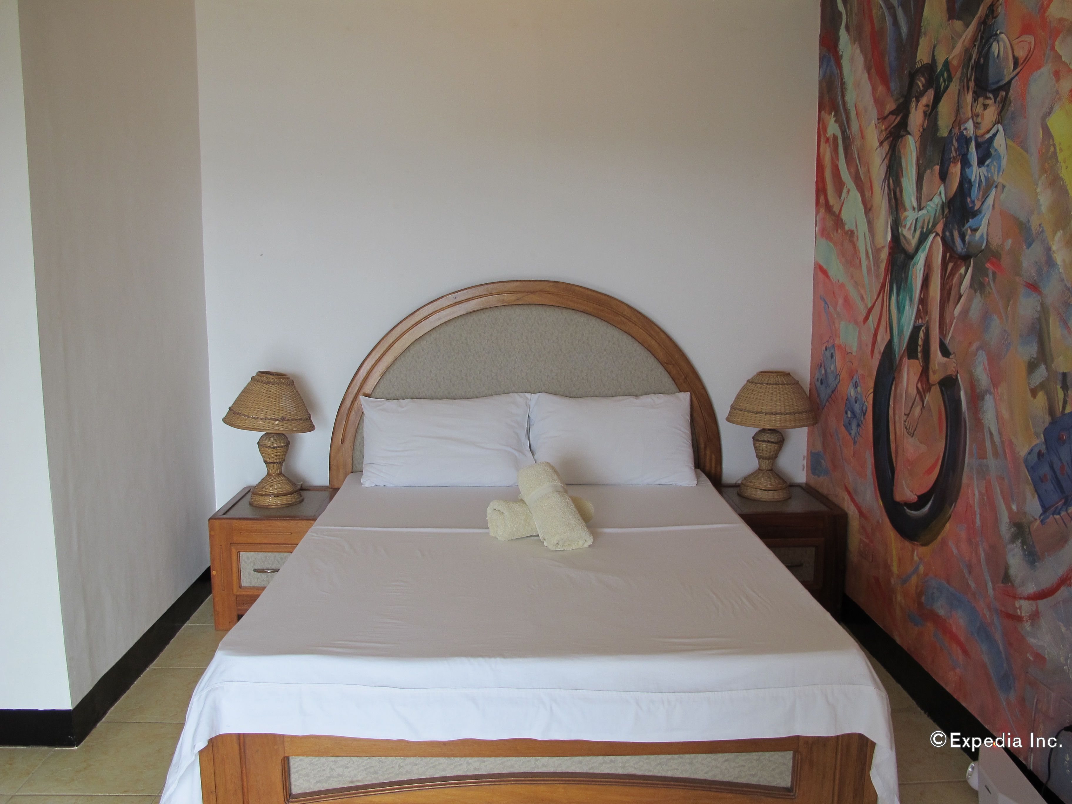 Una cama con cabecera de madera, ropa de cama blanca, dos lámparas de noche y un cuadro de colores en la pared.