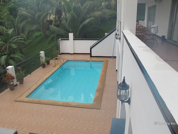 A rectangular swimming pool with a tiled deck, surrounded by a white wall and a balcony railing.