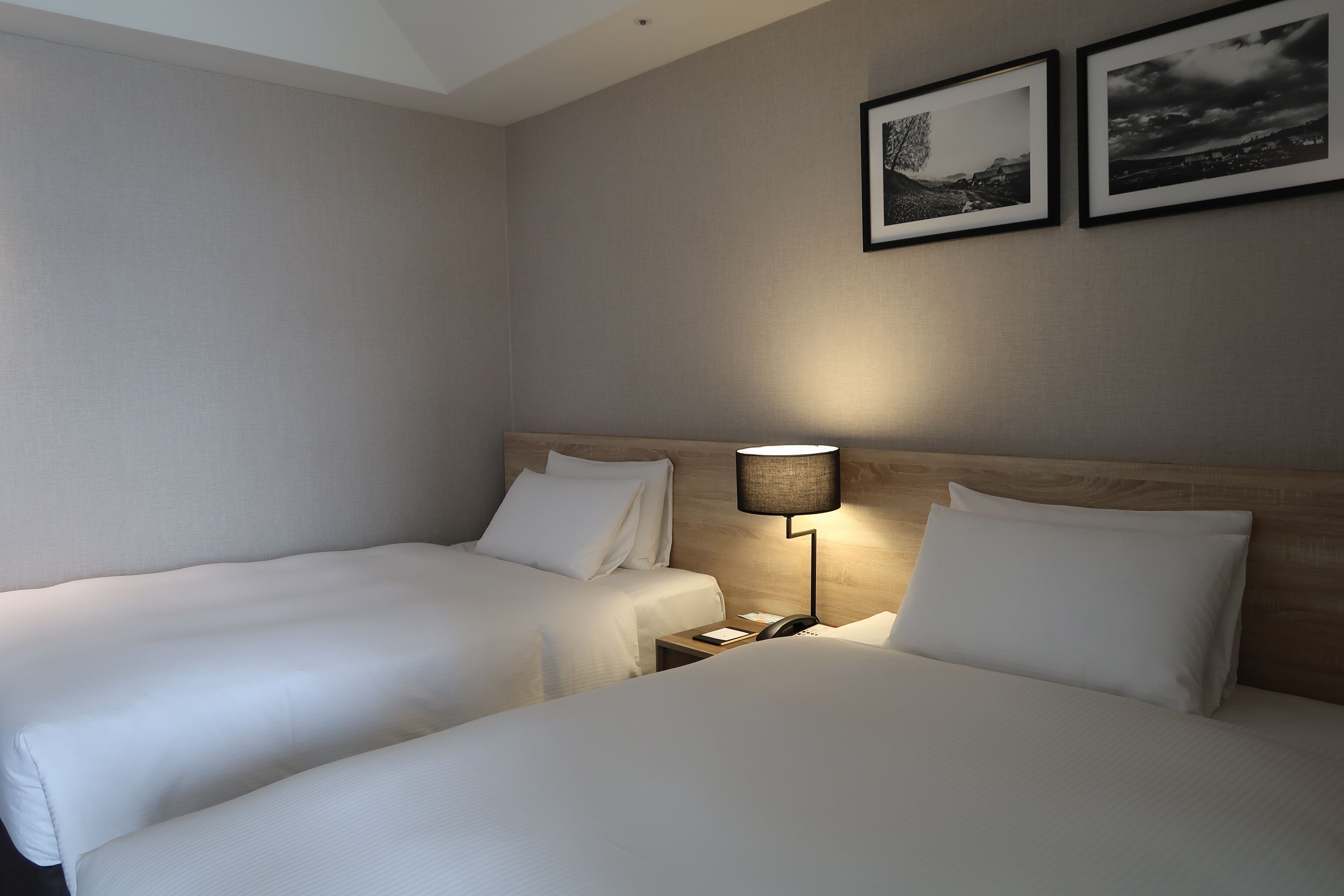 A hotel room with two beds, a bedside table, a lamp, and two framed pictures on the wall.