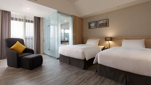 Een hotelkamer met twee bedden, een bank met een geel kussen, een nachtkastje met een lamp en uitzicht door een glazen deur.