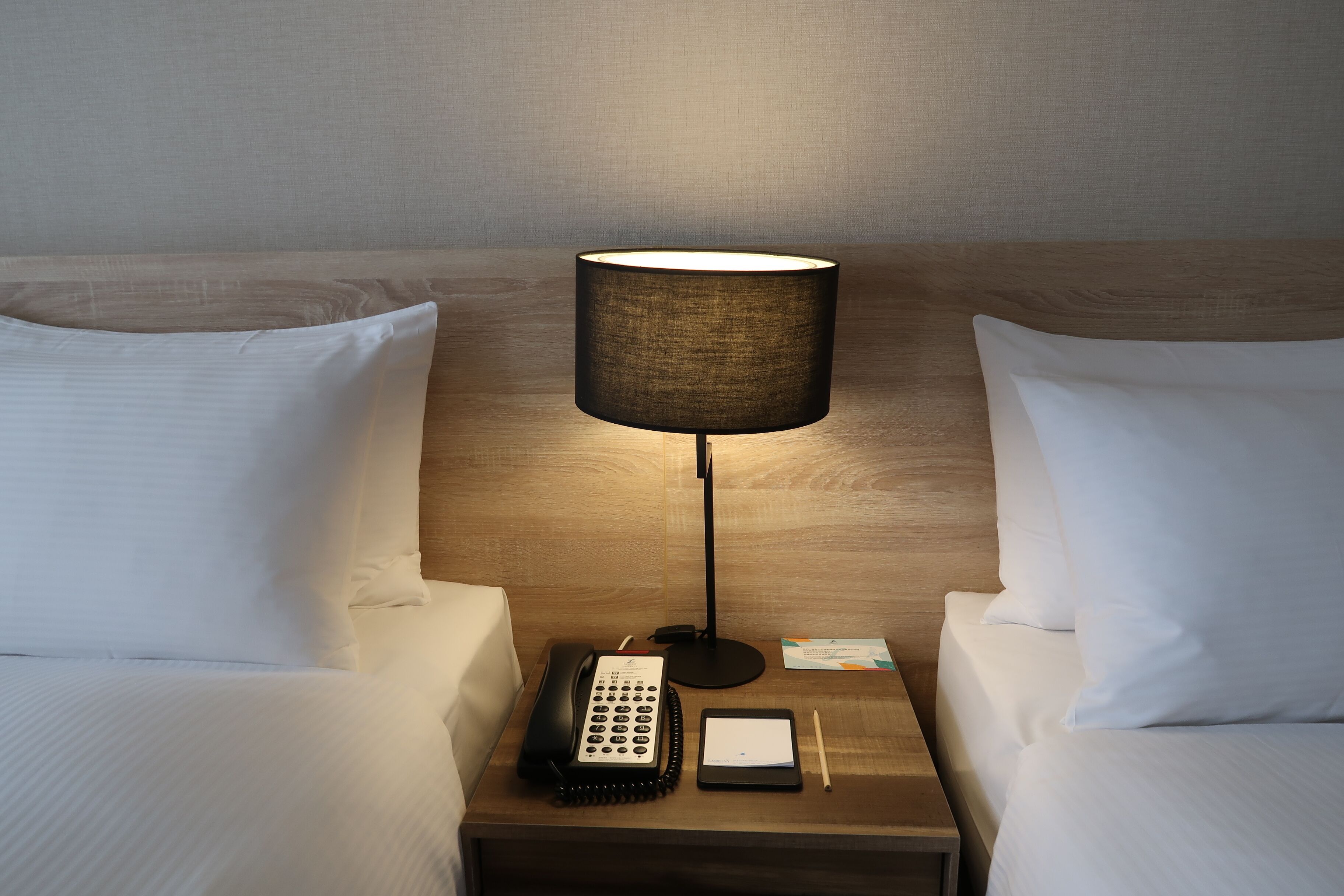 A hotel room with two beds, a bedside table with a lamp, a telephone, and a notepad.