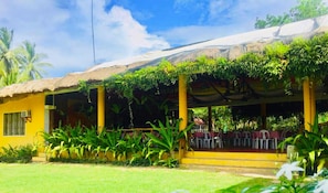 A yellow building with a thatched roof, covered outdoor seating area, and lush greenery.