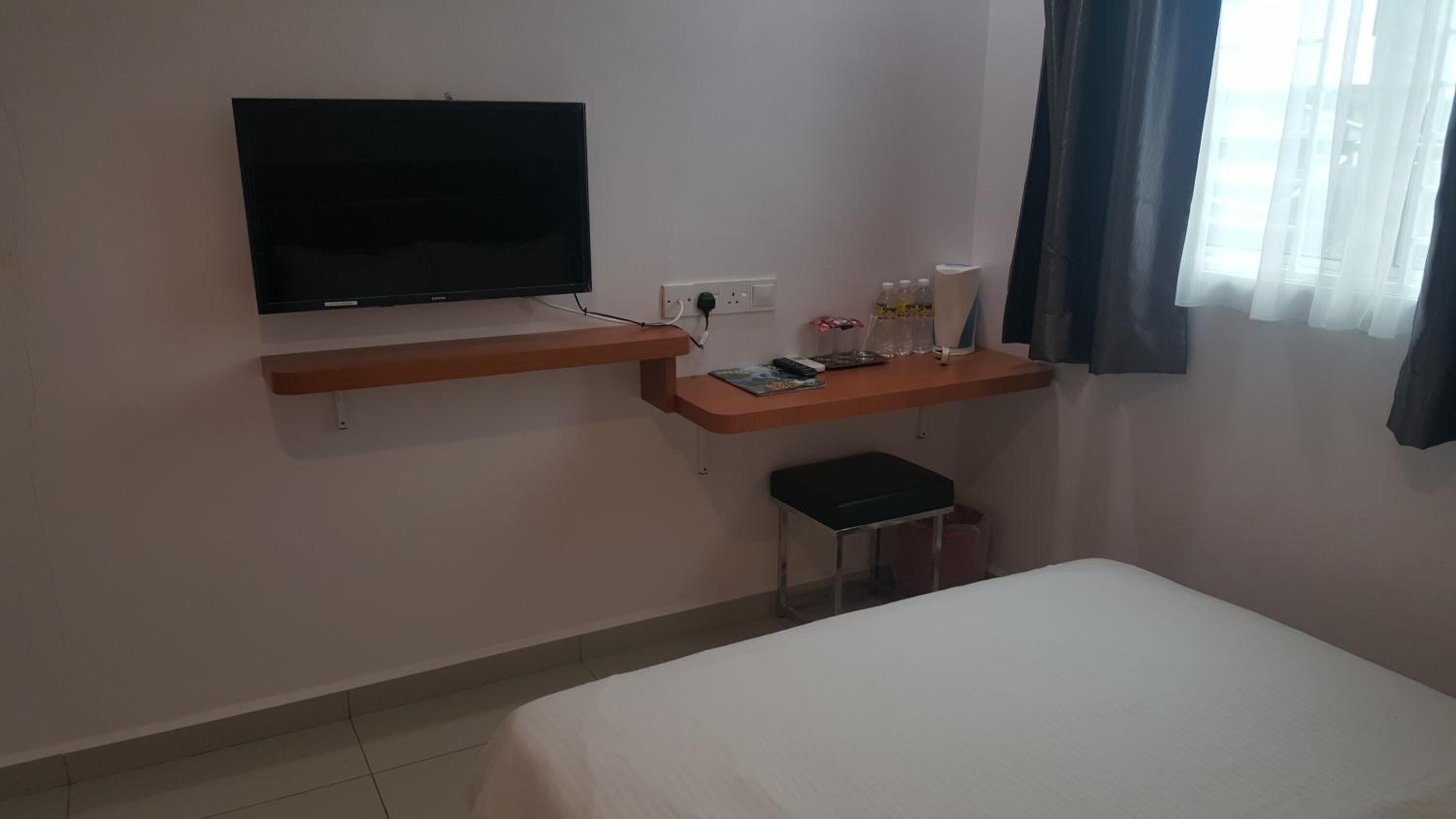 Habitación de hotel con cama, televisor montado en la pared, una mesita de noche con lámpara y ventana con cortinas.
