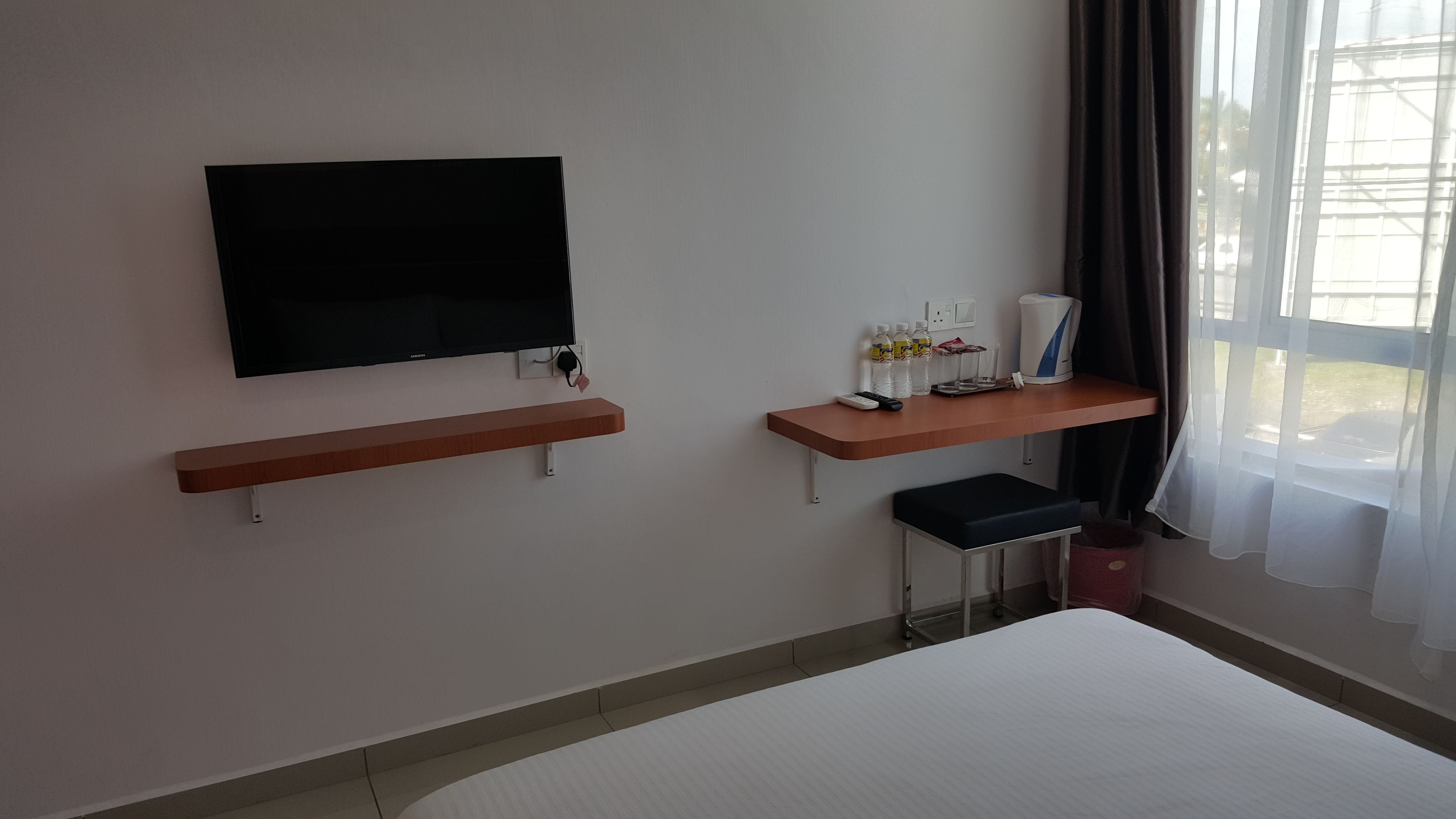 A hotel room with a flat-screen TV mounted on the wall, a wooden shelf with a remote control, a small table with a chair, and a bed.
