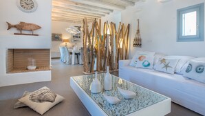 Un salón moderno con un sofá blanco, cojines decorativos, una mesa de centro de cristal con conchas marinas, un pez de madera en la pared y una estructura de madera rústica.