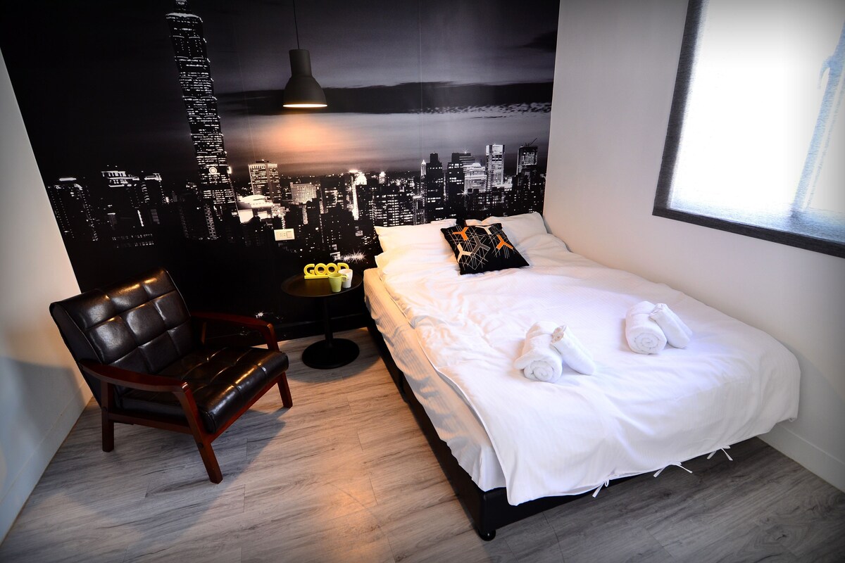 A modern hotel room with a cityscape mural, a bed with white linens, a leather chair, and a small side table.