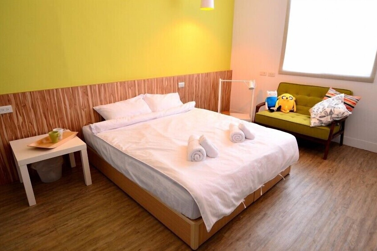 A hotel room with a wooden bed, a small bedside table with a cup, a green sofa, and a window with a view.