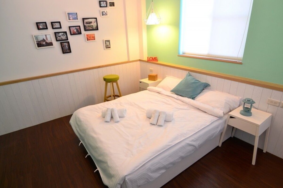 A neatly made bed with white linens and two towels, a bedside table with a lamp, a stool, and a wall with framed pictures.