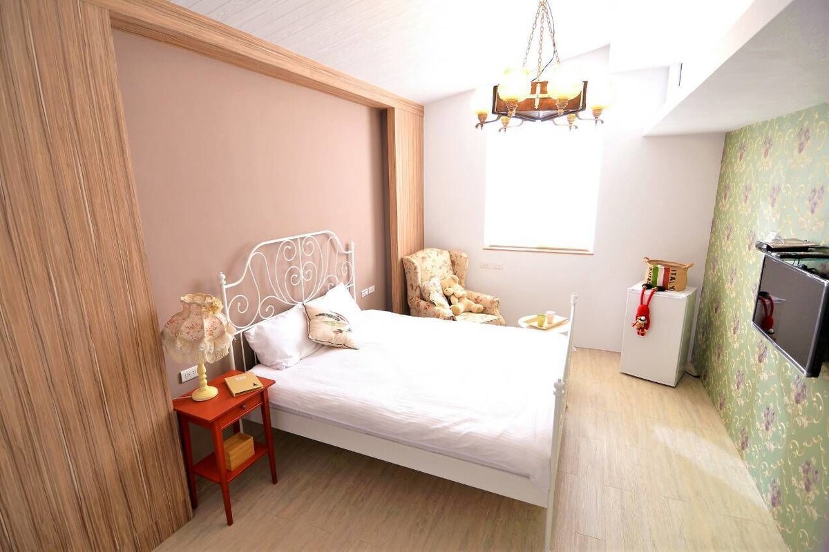 A bedroom with a bed, a nightstand, a chair, a desk, a refrigerator, and a window.