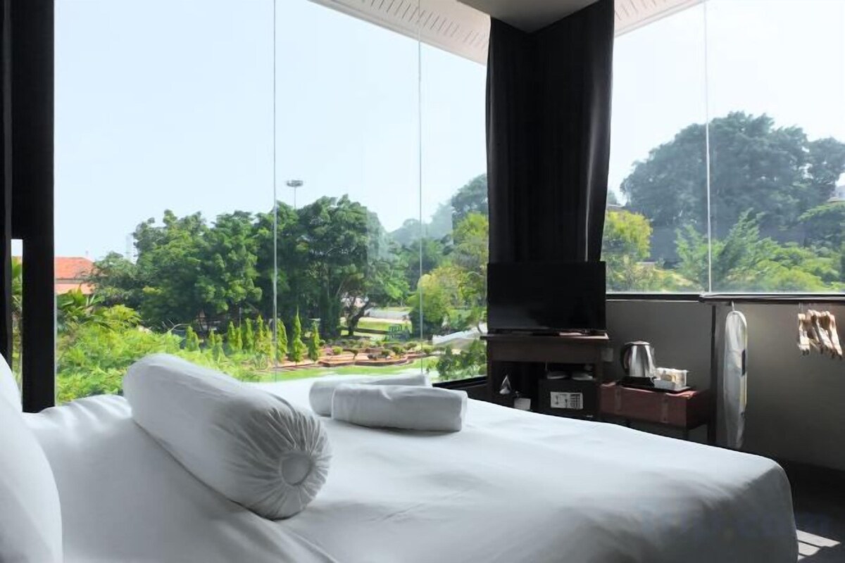 A hotel room with a bed, pillows, a television, a small table with a kettle, and a view of a garden and trees.