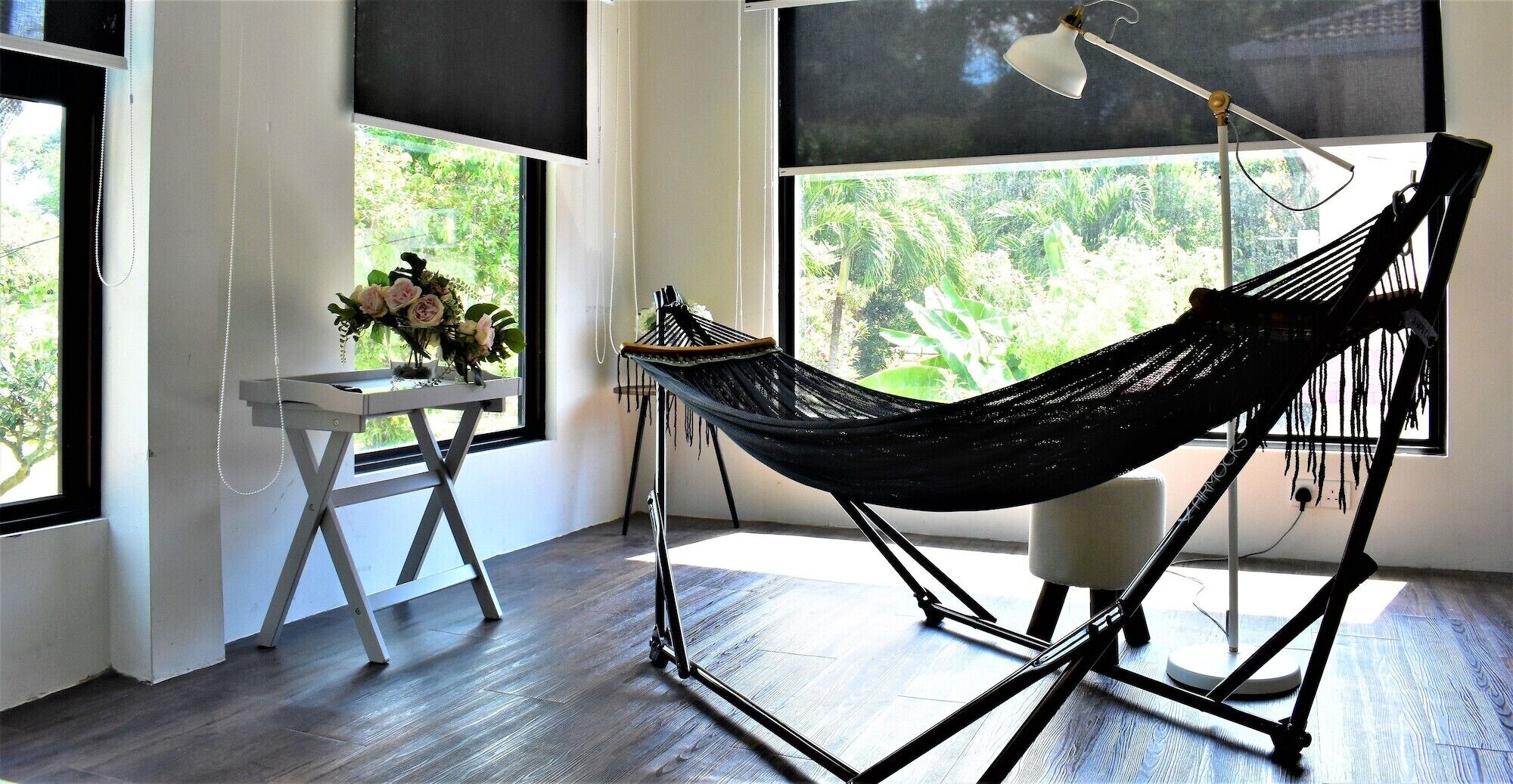 A hammock chair in a room with large windows, a small table with a vase of flowers, and a floor lamp.