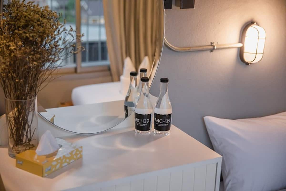 Une chambre d’hôtel avec une table de chevet, un vase contenant des fleurs séchées, deux bouteilles étiquetées « ARCH39 », une boîte de mouchoirs et un luminaire fixé au mur.