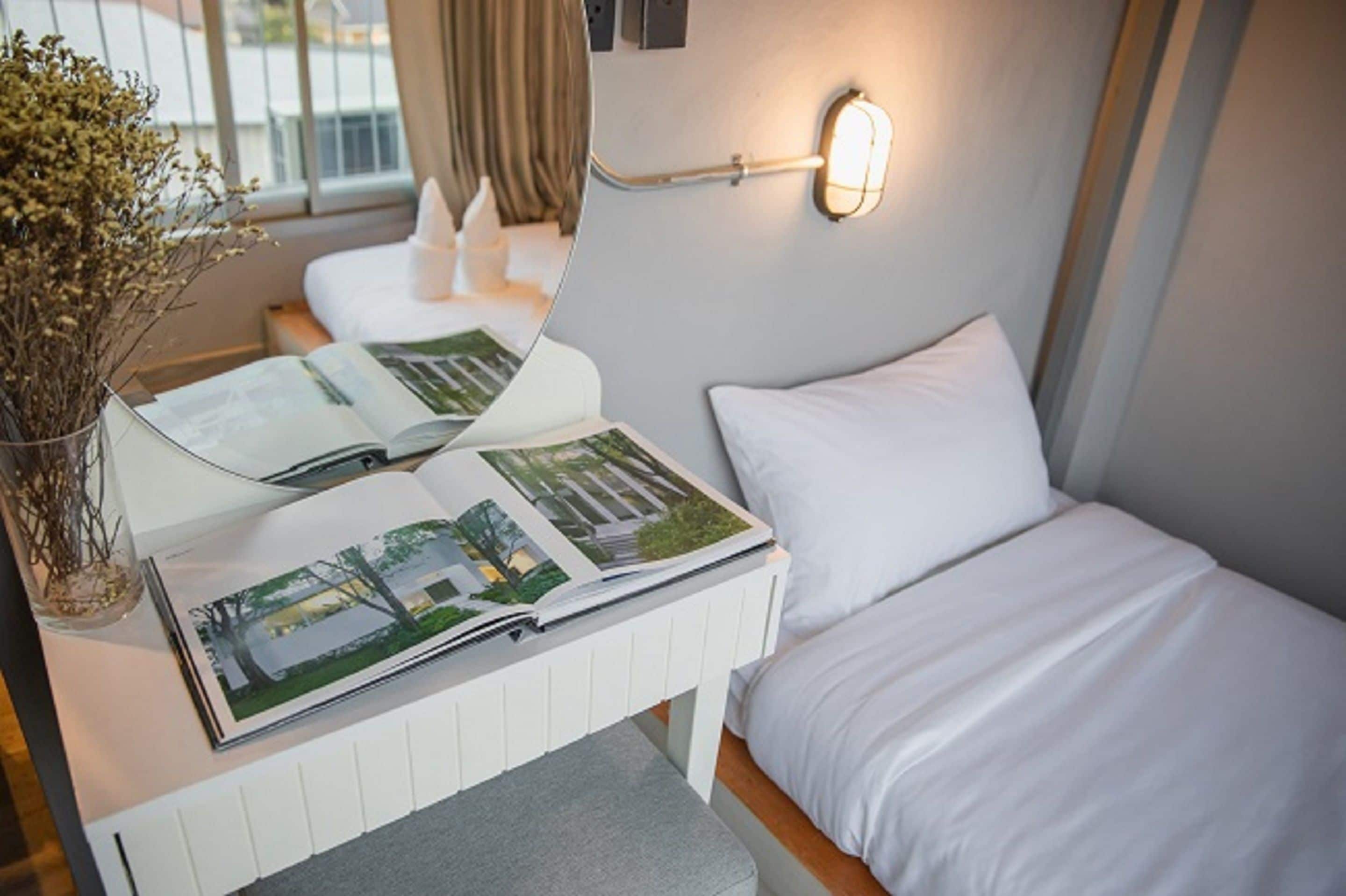 Une chambre d’hôtel avec un lit, une table de chevet sur laquelle se trouve un magazine, un vase avec des fleurs séchées et un luminaire fixé au mur.
