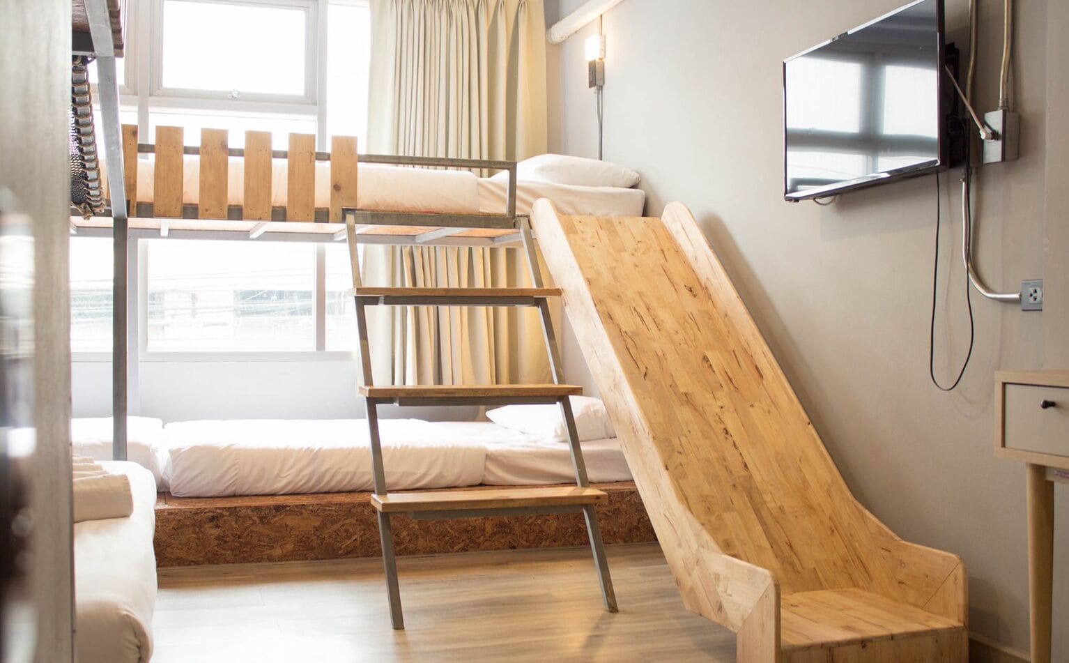 A bunk bed with a wooden ladder, a wooden slide, a television mounted on the wall, and a window with curtains.