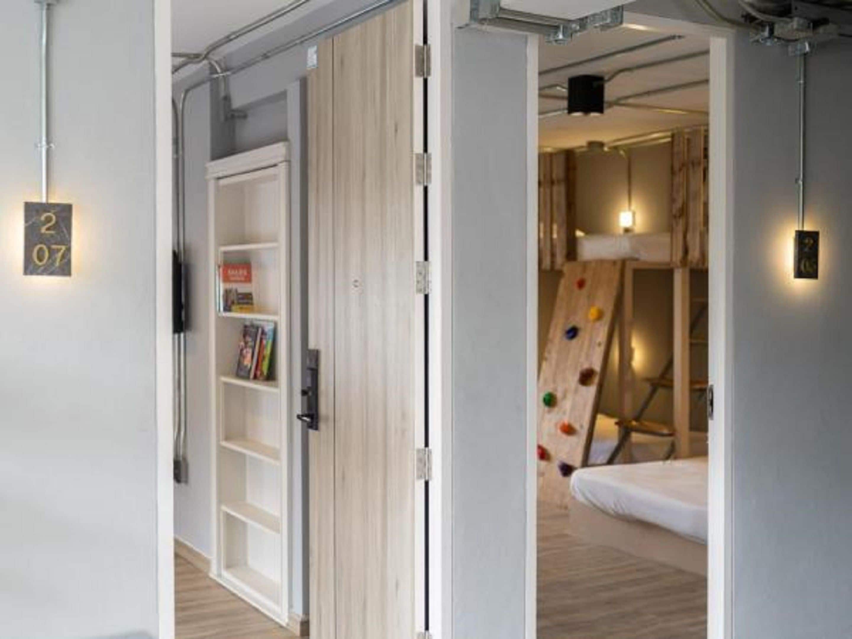 Una habitación moderna con una puerta blanca, una estantería y una cama con una pared de escalada al fondo.