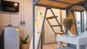 Una habitación con televisor montado en la pared, un refrigerador pequeño, una pared de escalar, un espejo redondo y una planta en maceta.