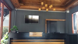 A modern reception desk with a wooden finish, a flat-screen TV mounted above it, and a large mirror to the right. - Le Terrarium Hotel (Chiang Rai)