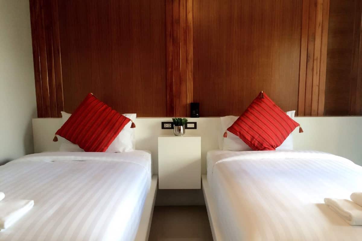 A hotel room with two beds, each with white linens and red pillows. There is a small table with a plant and a clock between the beds.