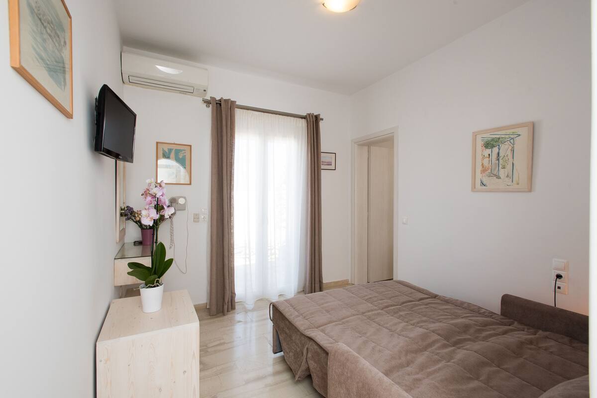 A bedroom with a bed, a bedside table with a potted plant, a flat-screen TV mounted on the wall, and a door leading to another room.