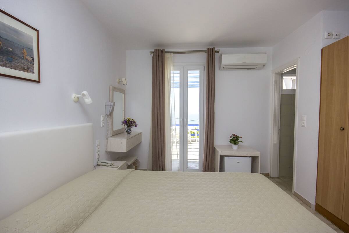 A bedroom with a bed, a bedside table with a vase, a mirror, a window with curtains, and an air conditioning unit.