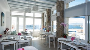 A bright dining area with white furniture, large windows, and a view of the sea. - Spanelis Hotel (Mykonos)