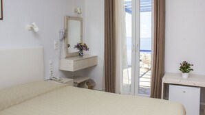 A hotel room with a bed, a small desk, a mirror, a telephone, a plant, and a view of the sea through a sliding door. - Spanelis Hotel (Mykonos)
