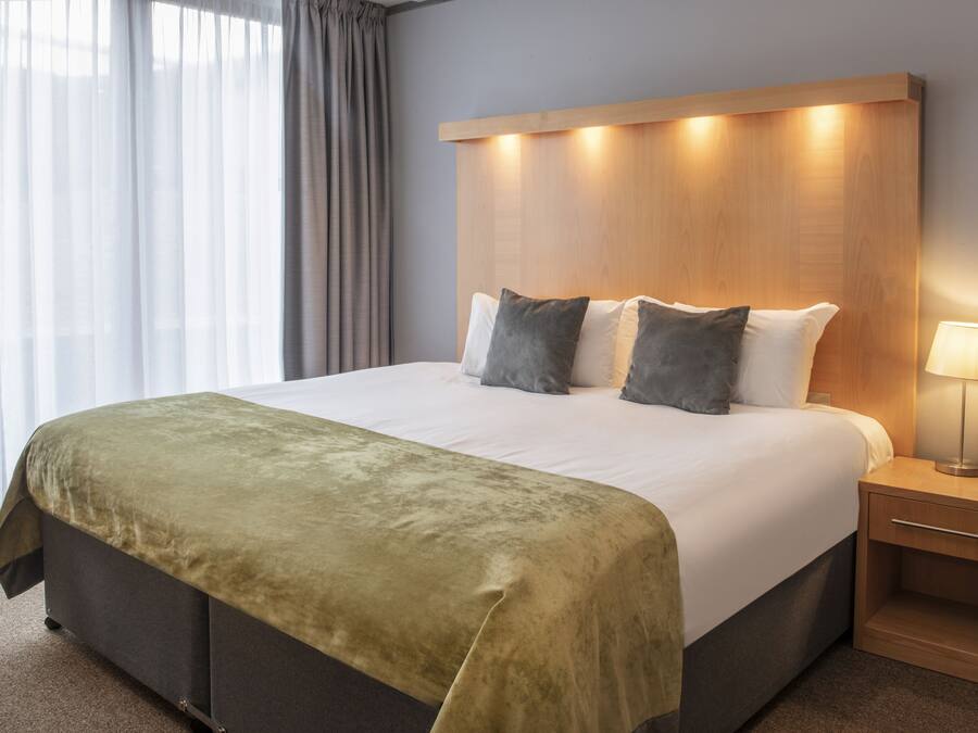 A hotel room with a large bed, a bedside table with a lamp, and a view through sheer curtains.