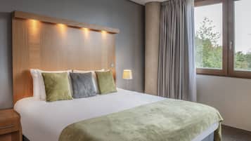 A hotel room with a large bed, a wooden headboard with built-in lighting, a bedside lamp, and a view of trees through a window with curtains.