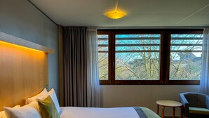 A hotel room with a large bed, a small round table, a chair, and a window with a view of trees and buildings. - The Jubilee Hotel & Conferences (Nottingham)