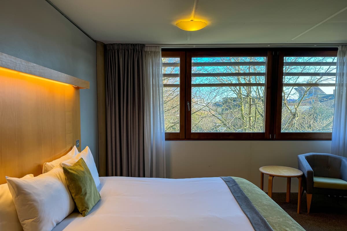 A hotel room with a large bed, a small round table, a chair, and a window with a view of trees and buildings.