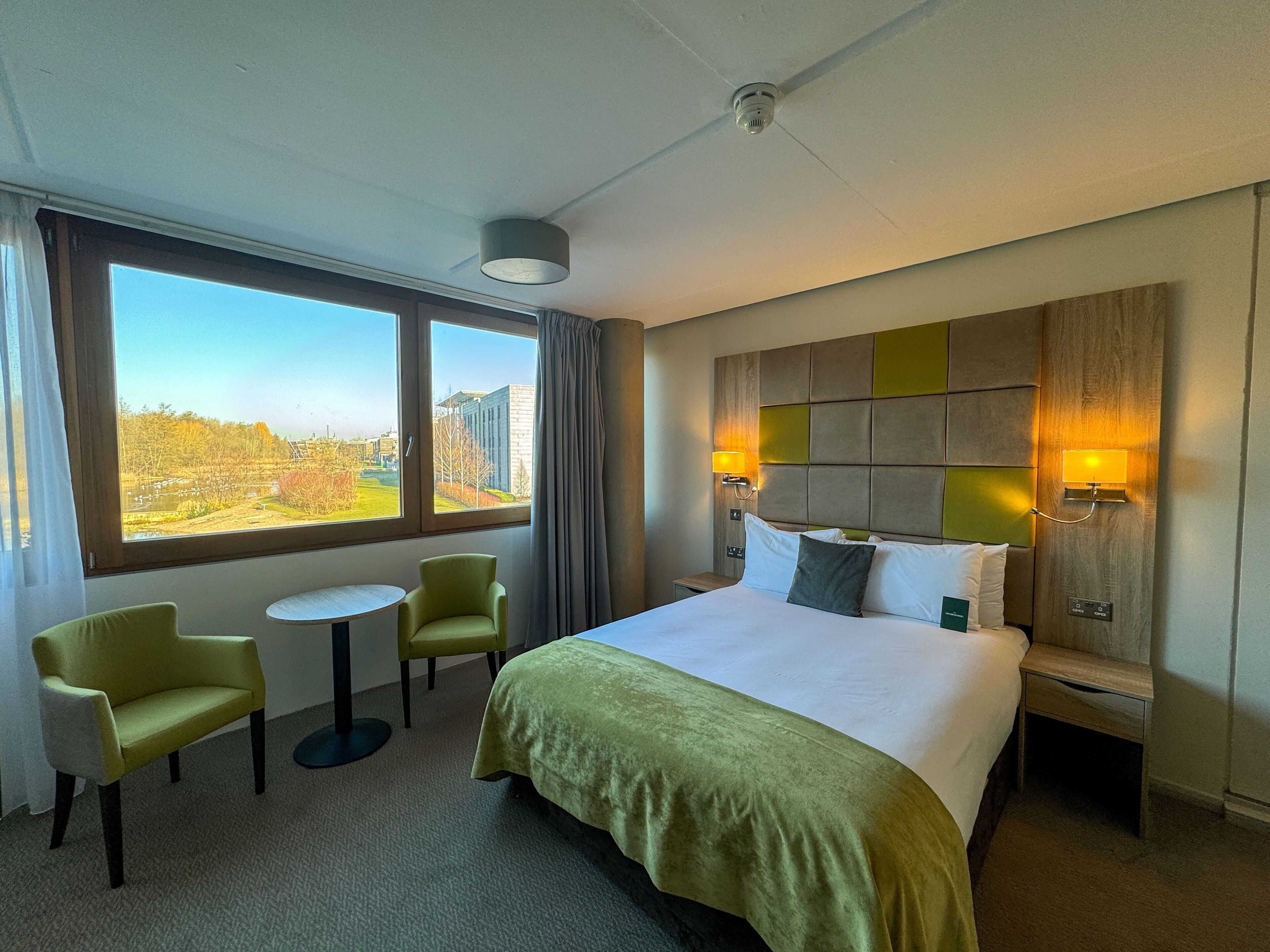 Ein Hotelzimmer mit einem Bett, zwei Sessel, einem kleinen Tisch, einem Nachttisch und Blick auf ein Gebäude und Bäume durch große Fenster.