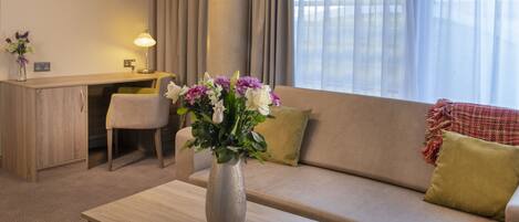 A modern hotel room with a sofa, a small table, a desk, and a window with curtains.