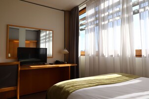 A hotel room with a bed, a desk with a television, a mirror, and a window with curtains.