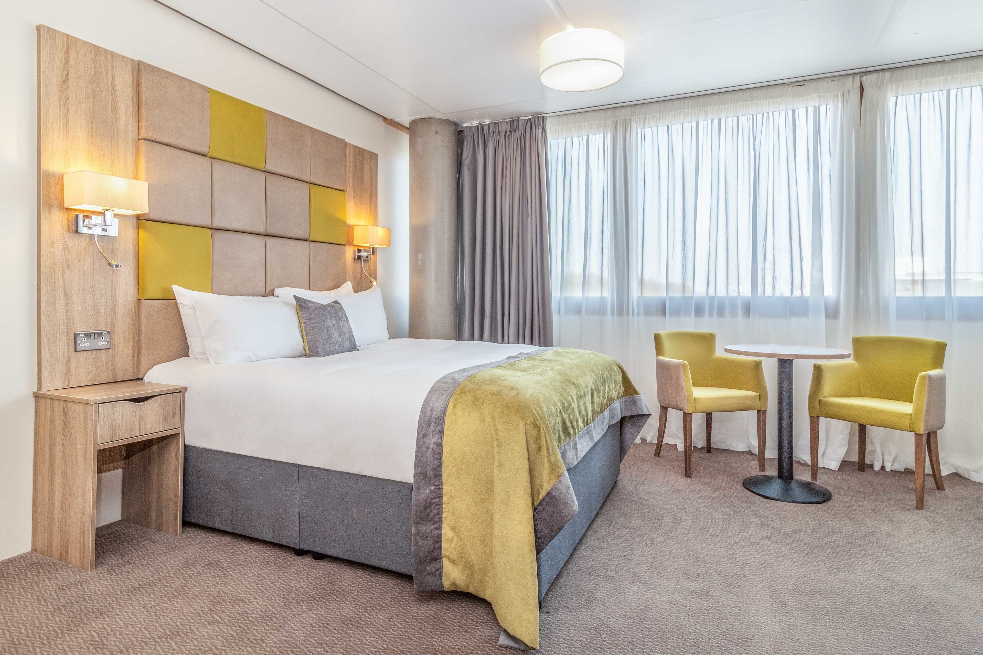 A hotel room with a large bed, bedside tables, a round table with two chairs, and a view of the outdoors through large windows.