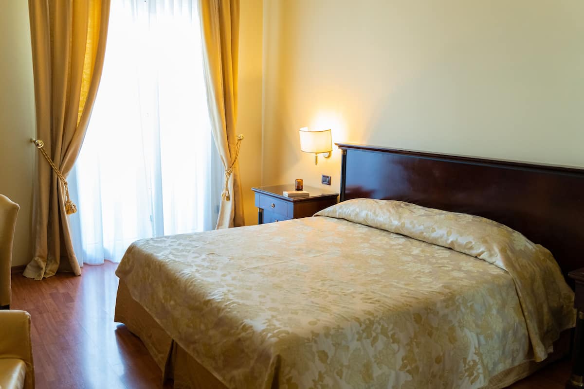 Een hotelkamer met een groot bed, een nachtkastje met een lamp en een raam met gordijnen.
