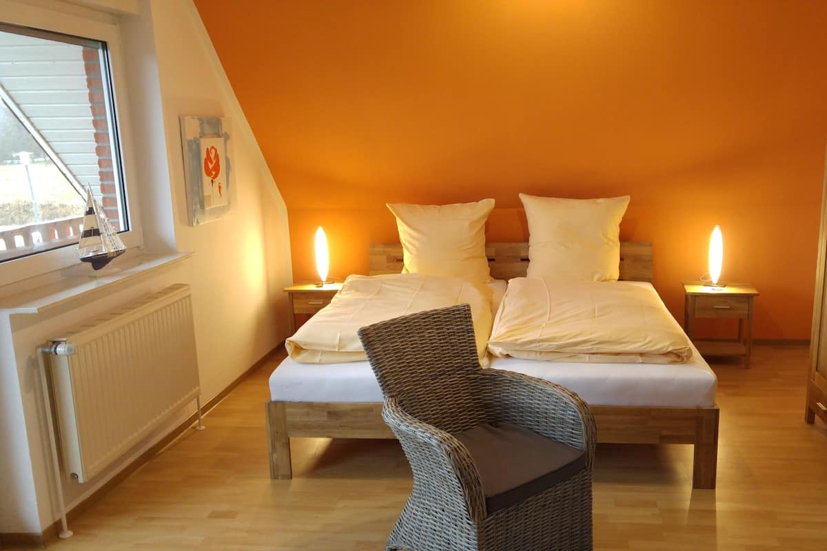 A bedroom with a bed, two bedside tables with lamps, a wicker chair, a wooden wardrobe, and a window with a sailboat decoration.