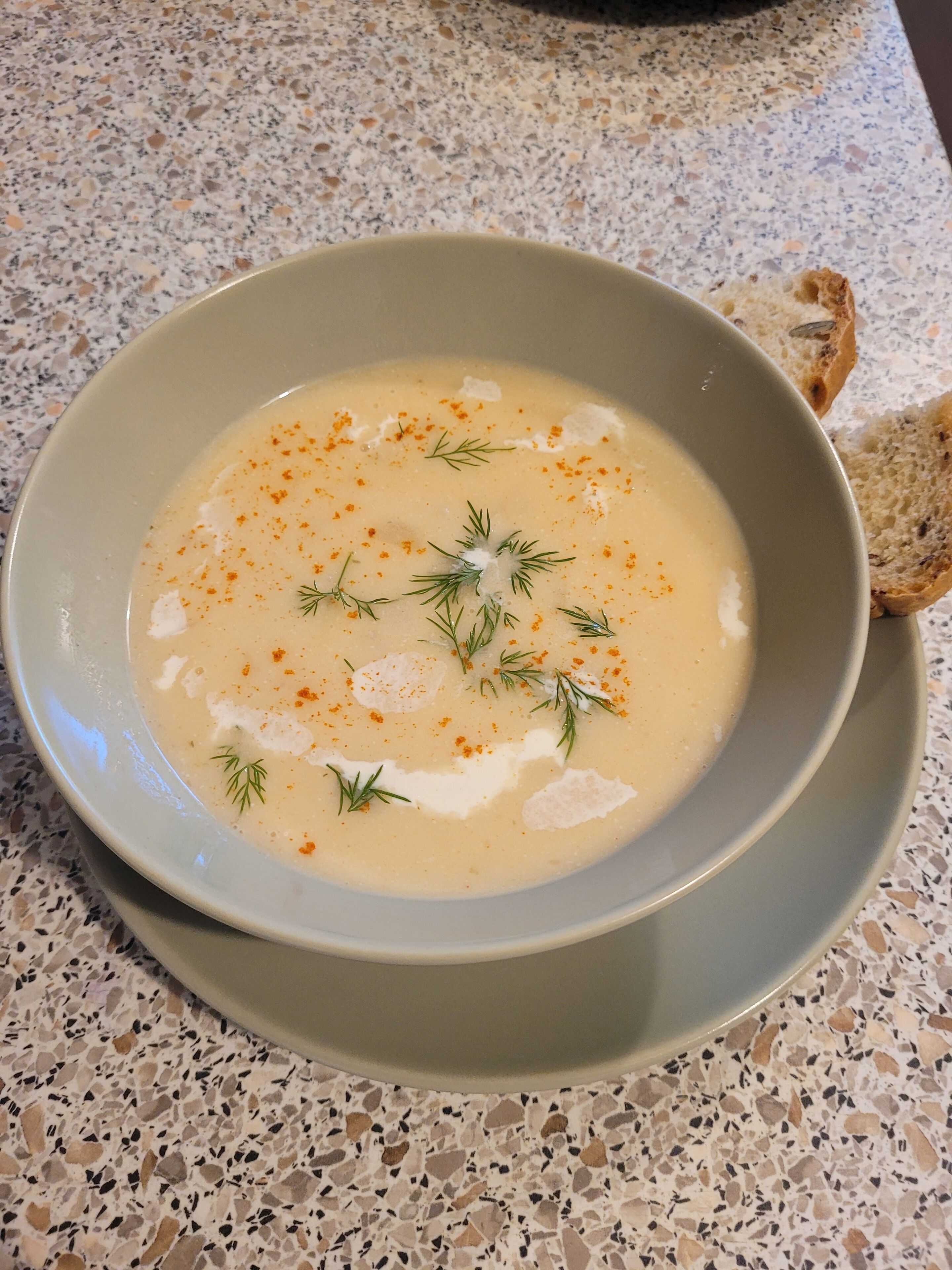 A bowl of creamy soup garnished with dill and a sprinkle of red spice, served on a speckled countertop.