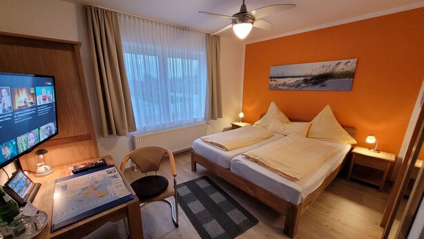 Una habitación de hotel con cama, escritorio con computadora, silla, ventana con cortinas, ventilador de techo y un cuadro en la pared.
