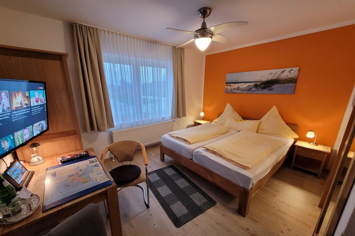 A hotel room with a bed, a desk with a computer, a chair, a window with curtains, a ceiling fan, and a painting on the wall.