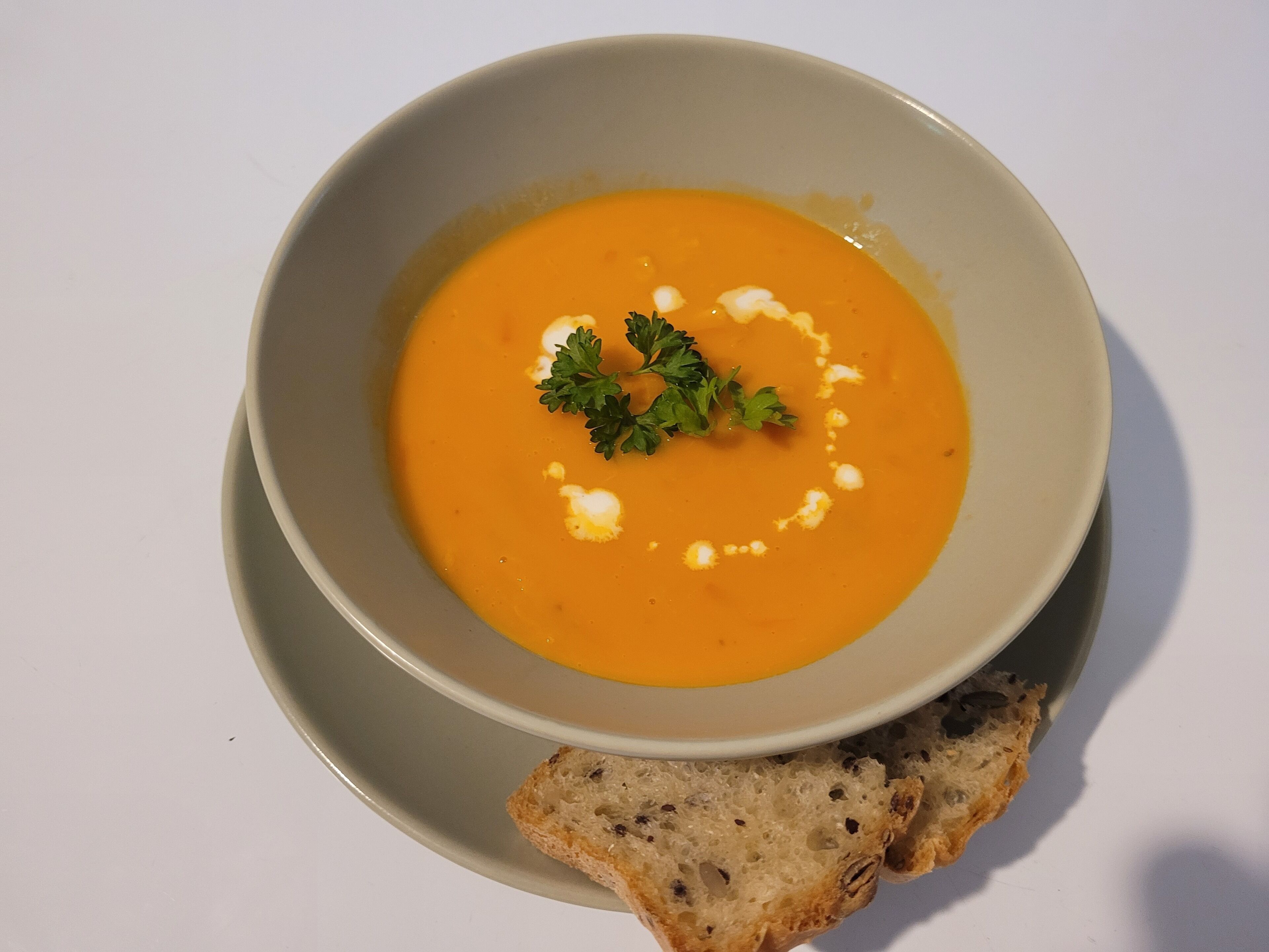 Un bol de soupe agrémenté d’une garniture, servi avec une tranche de pain sur une assiette.