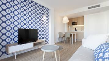 A modern living room with a flat-screen TV, a grey round table, and a sofa. There is a kitchen area with wooden cabinets and a dining table with chairs.