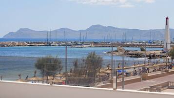 Una marina costera con barcos, un faro y montañas al fondo.