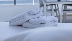 Ein ordentlich gefalteter Stapel weißer Handtücher auf einem Bett mit Blick auf das Meer und einem Essbereich im Hintergrund.