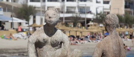 A beach with two stone sculptures, a white building with balconies, and people in the background.