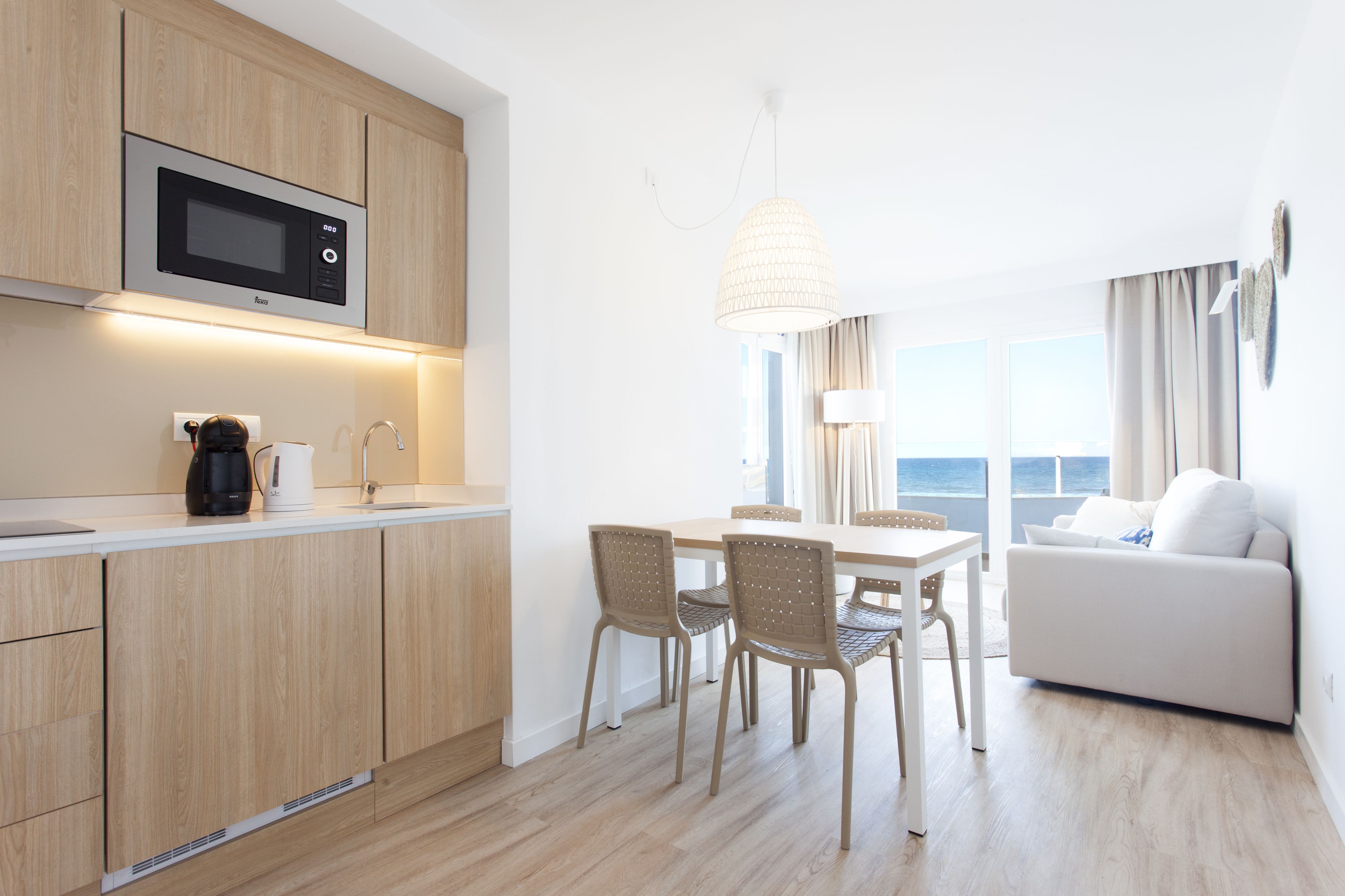 Una cocina moderna con armarios de madera, microondas integrado y cafetera. Hay una zona de comedor con mesa y sillas, y un sofá blanco en la sala de estar.