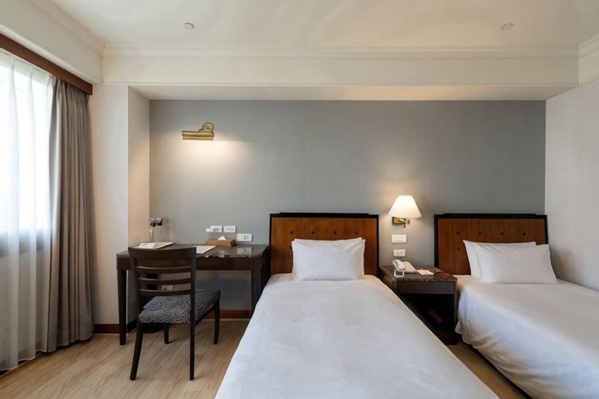 A hotel room with two beds, a desk, a chair, and a nightstand with a lamp.
