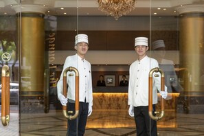 Two waitstaff holding large, ornate keys in front of a hotel reception area with a marble floor and a chandelier.