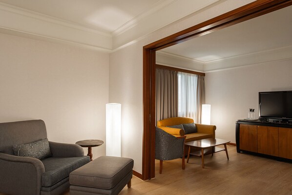 A modern hotel room with a grey sofa, a small round table, a wooden coffee table, a yellow armchair, and a television on a wooden cabinet.
