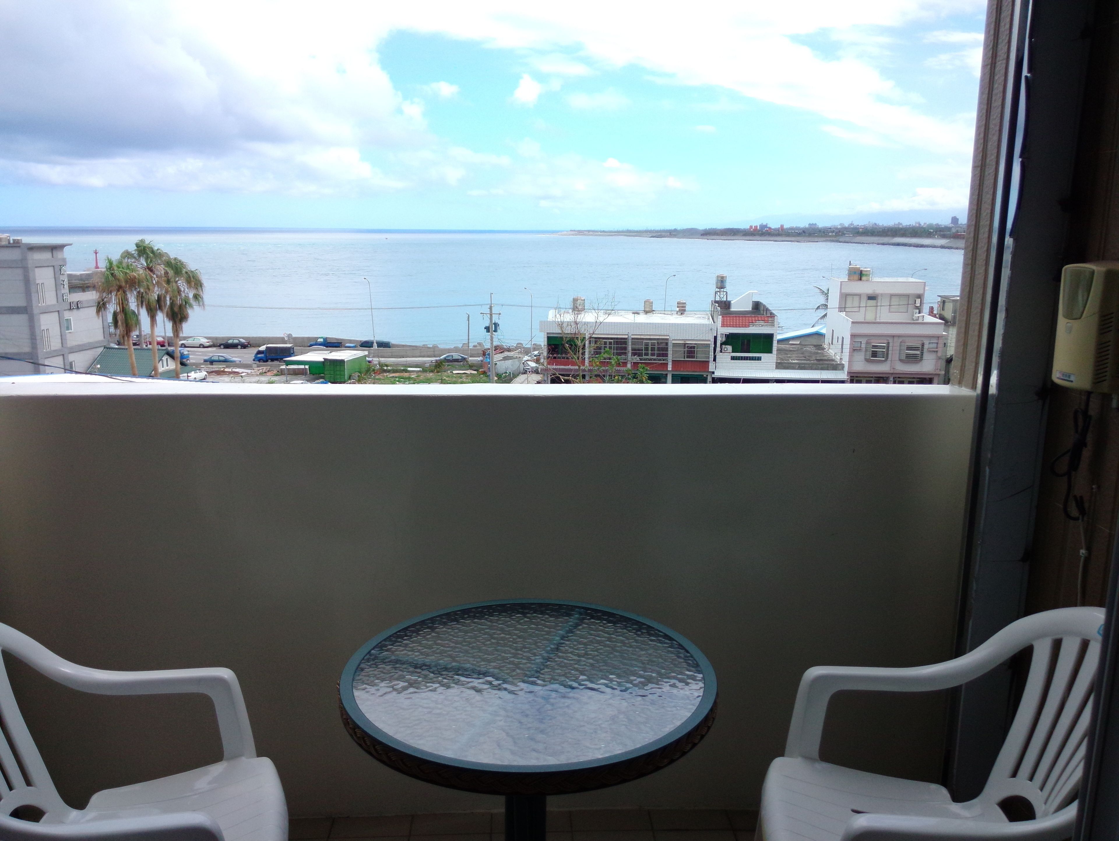 A balcony with a glass table and two chairs, overlooking a coastal area with buildings and palm trees.